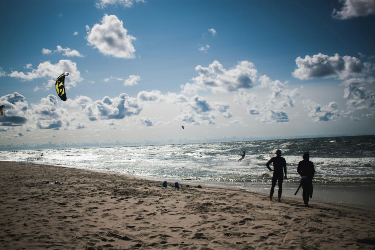 Two people watching kite surfers at sunny, cloudy beach